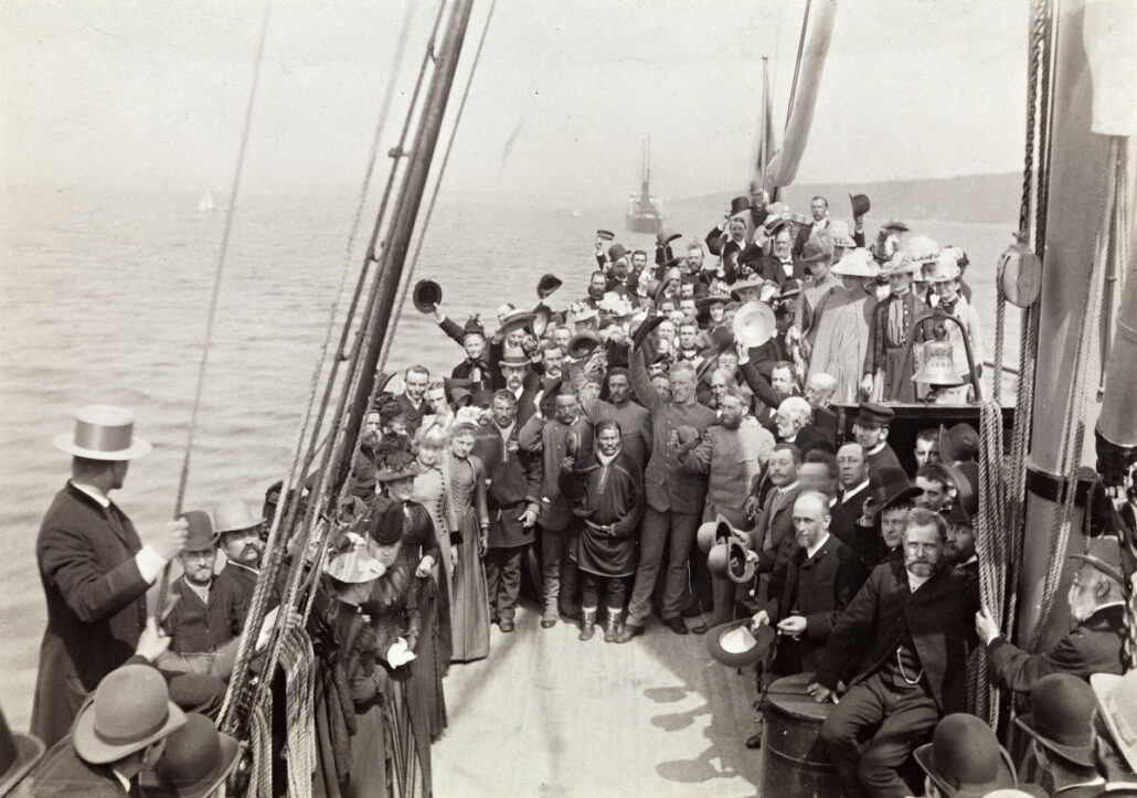 Gruppebilde av ca 5o glade og festkledde personer på dekket av en båt, man ge holder hatten i været