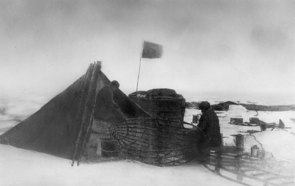 Teltleir på sneen, et flagg vaier utenfor teltåpningen og to personer står ved inngangen og arbeider