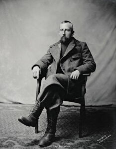 Portrett av mann i ulldress, han sitter i stol med armene på armlen og venstre ben krysset over det høyre.