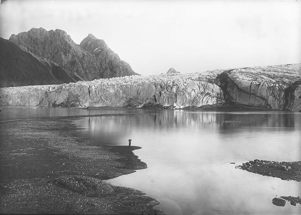 Stor isbrefront, i forgrunnen en fjord med en liten skikkelse.