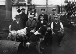 Gruppebilde av åtte menn på båtsdekk sammen med to store hunder
