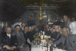 23 menn sitter/står rundt et bord med dramglass og flasker i et lite intimt rom med skråtak og parafinlampe i taket.