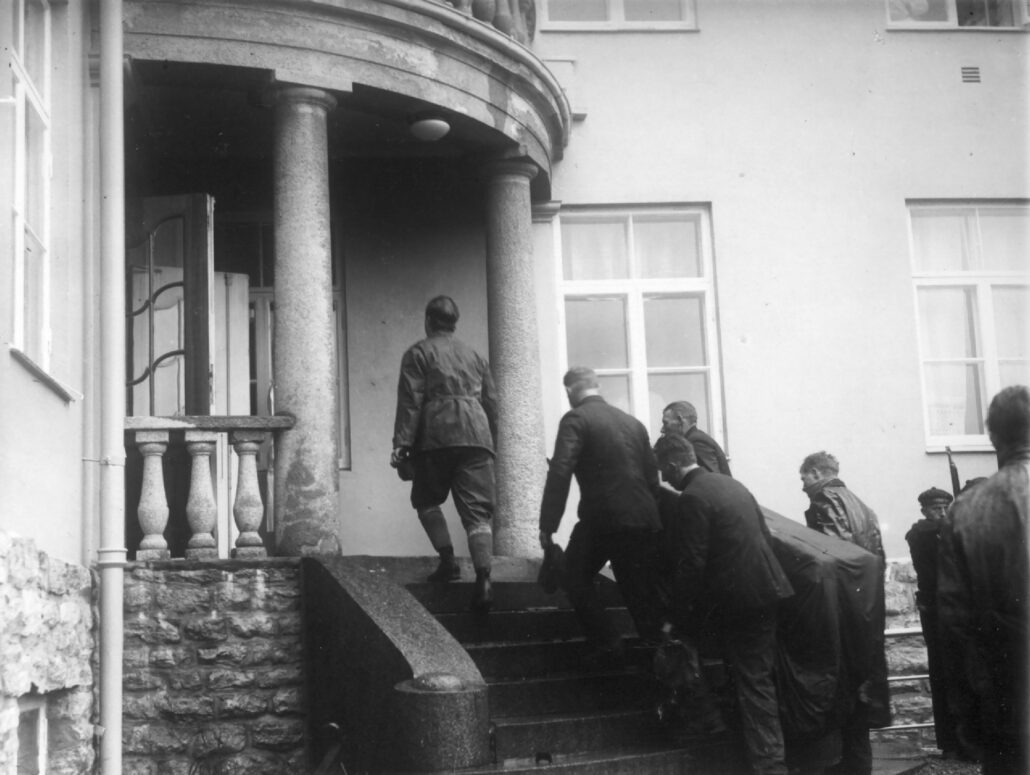 Fire menn bærer en smal og høy kasse dekket med et sort klede opp en trapp og inn en bygning