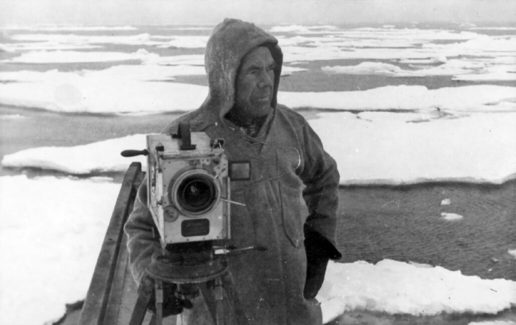 Mann med videokamera står på is med hav i bakgrunnen.