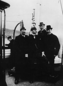 Fire menn poserer på dekket av en båt. De har mørke penklær, hatter og bart.