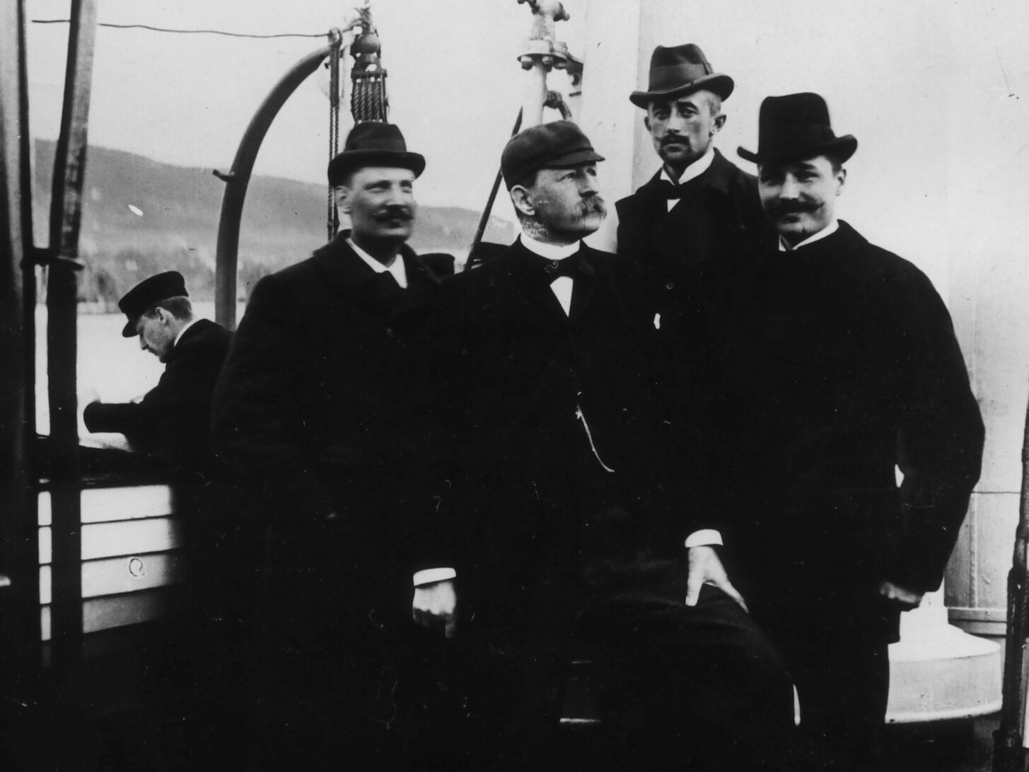 Fire menn poserer på dekket av en båt. De har mørke penklær, hatter og bart.