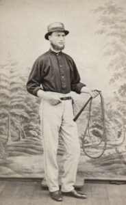 Mann i sort skorte, hvit bukse og hatt holder en pisk i venstre hånd. Bildet er tatt i studieo med et skog bilde som bakgrunn