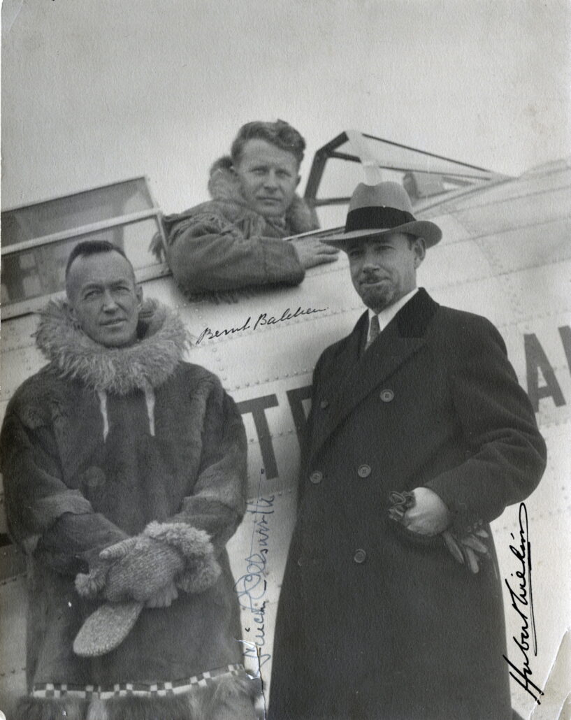 To menn står foran et lite fly, den ene har pelsklær og votter, den andre frakk og hatt. Bak dem sitter en mann i åpen cockpit på et lite fly. Under armen som hviler på flykroppen står det Bernt Balchen.