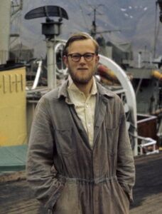 Mann med briller og skjegg i arbeidsoverall på en kai med et skip i bakgrunnen