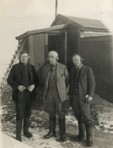 Tre menn står sammen foran en trebygning med en stige lent opptil veggen