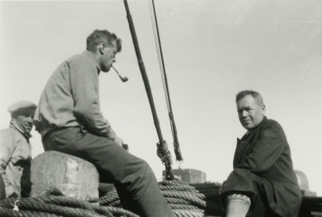 Tre menn står/sitter på et båtdekk. Det er mye tau kveilet opp. En røyker pipe
