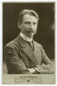 Portrett av ung mann med bart og flippskjegg. Han har ulldress og armene i kors