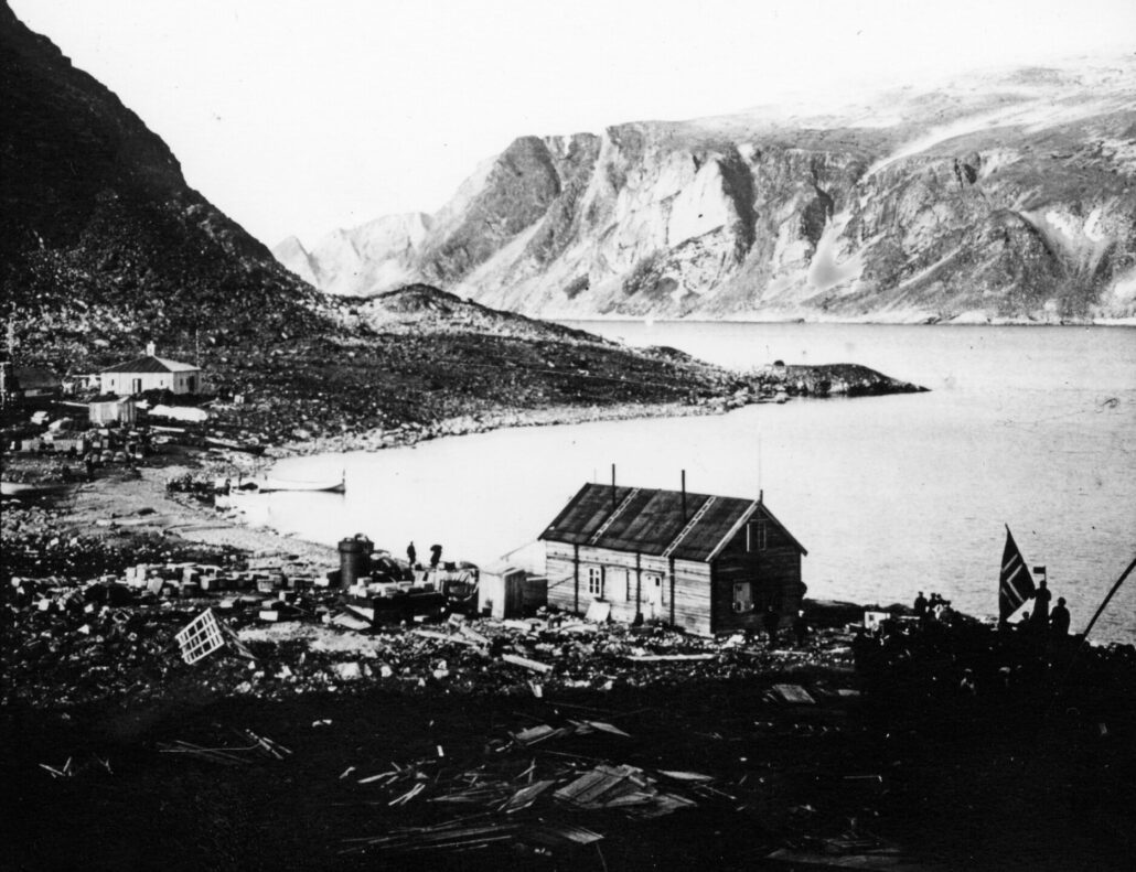 sort-hvit landskapsbilde med hus ved kysten. Fjell ligger til venstre og i bakgrunnnen