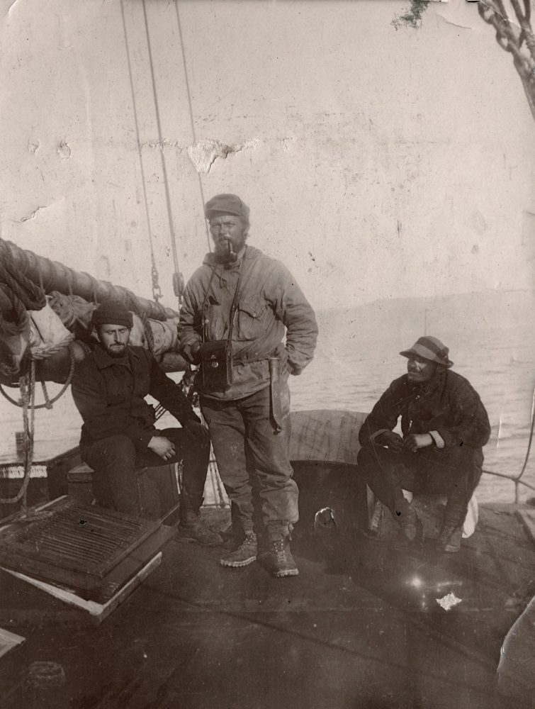 To menn istter og en står på et båtdekk. Han som står har pipe i munnen og en kikkertveske rundt halsen.