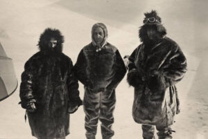 Tre personer i pelsklær med hette står på snedekt bakke