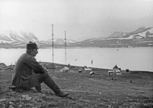 Mann sitter på toppen av en helning og skuer utover havet. Nederst ved bakken, mot havet er en klynge hus pg høye radiomaster