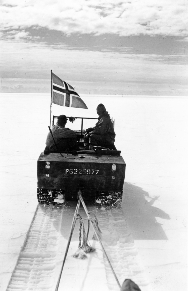 beltevogn kjører i snøen, to personer sitter, på norsk flagg vaier på venstre side