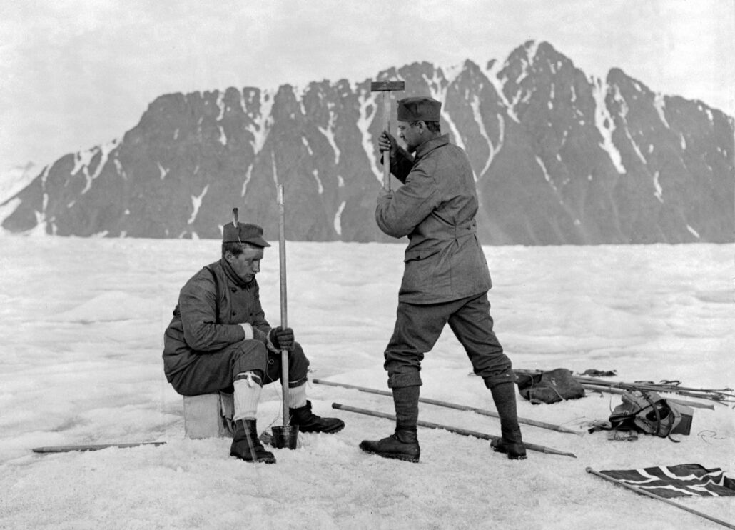 Sittende mann holder en pinne i isen, stående mann holder slegge og er klar til å slå pinnen. Rundt dem på snøen ligger utstyr og et flagg