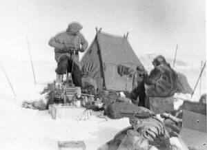 Mann sitter på ergometersykkel ute i snøen foran et telt og gire energi til en annen mann som bruker radio.