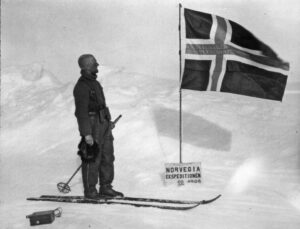 Mann på ski ser på det norsk flagg som vaier i snøen med et skilt under. Han holder lua i handa