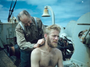 Mann klipper håre til en mann i bar overkropp på skipsdekk.