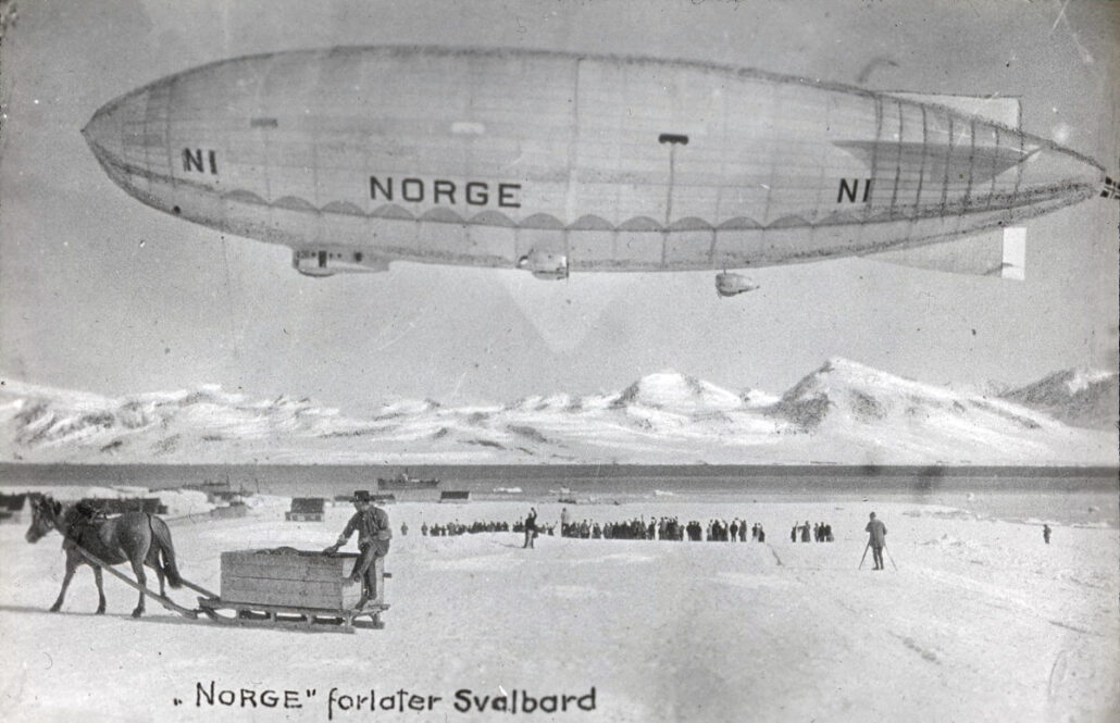 Luftskipt med påskrift Norge flyr over bakken hvor en gruppe mennesker står og ser på. En mann med hest og slede står i venstre forkant og det er snedekte fjell bak