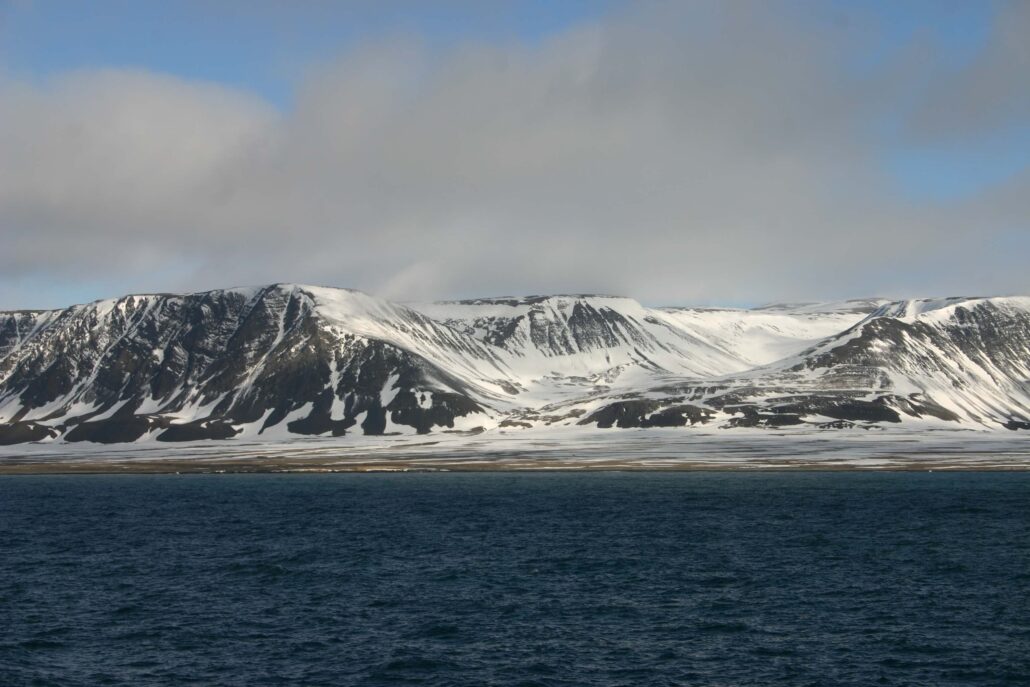 landskapsbilde med snødekkede fjell ved kysten. Hav er mørk blå og himmelen er litt overskyet.