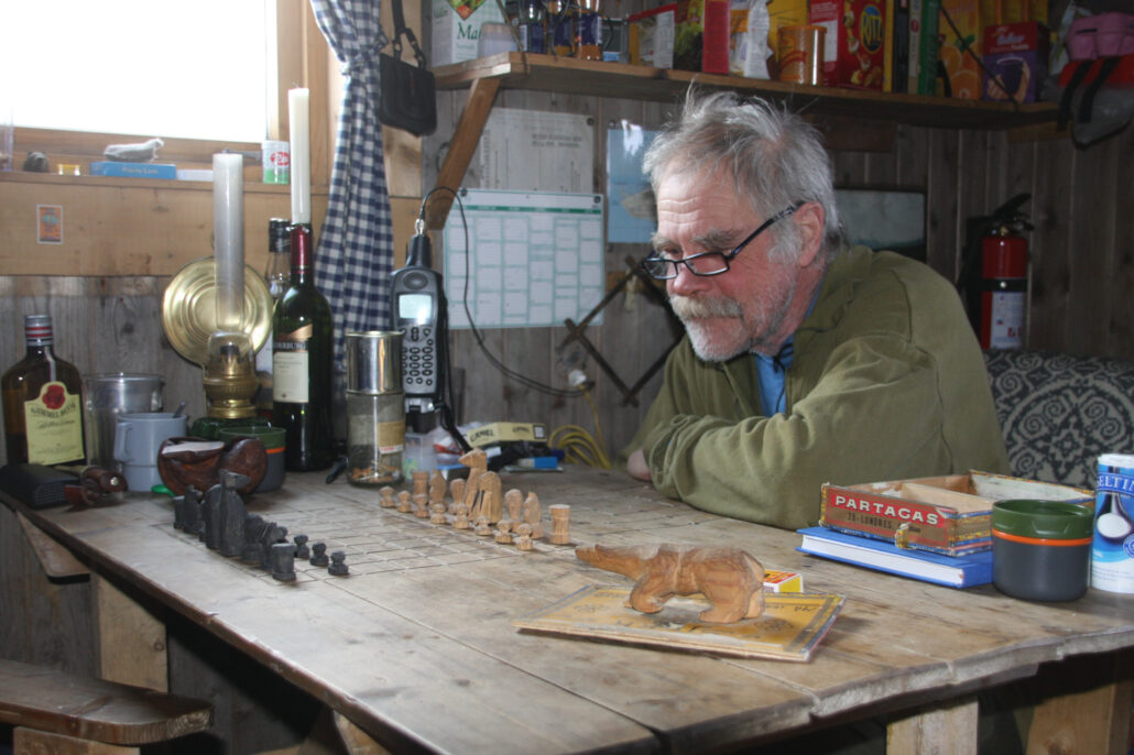 Eldre mann med briller sitter ved en bord med hjemmelagde sjakkbrikker og sjakkbrett risset inn i bordplaten