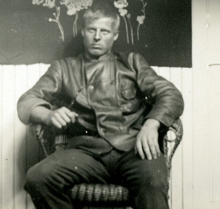 Mann i skinnjakke sitter i en kurvstol, en dør til hans høyre og et blomsterbilde på veggen bak han