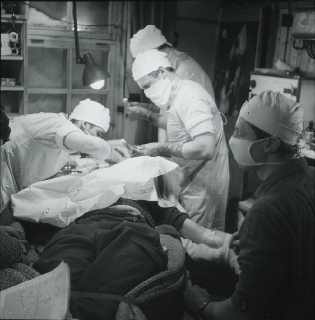 Mann ligger på operasjonsbord med hverdagsklær, rundt står og sitter fire personer med operasjonsklær