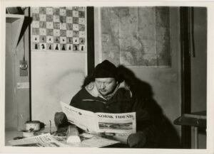 Mann leser avis med vinterklær og votter på, inne ved et bord, bak henger et kart og et sjakkbrett i tekstil