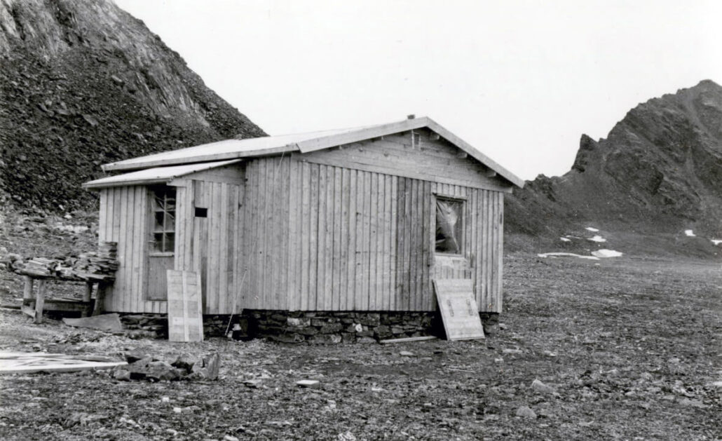 sort-hvit foto som viser ei hytta i tre. Fjell ligger i bakgrunnen.