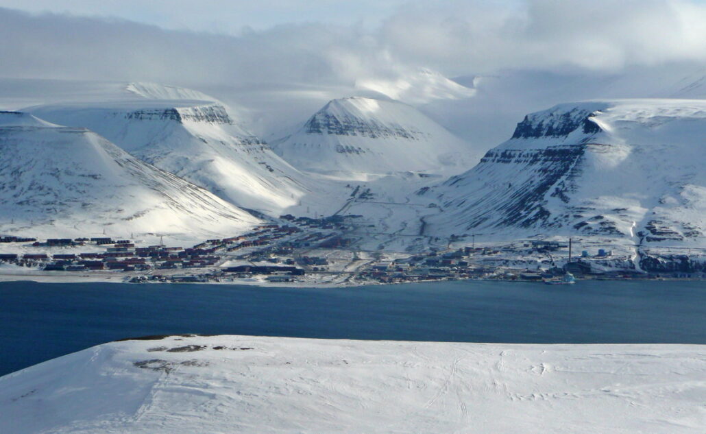 Bebyggelse i en dal ved en fjord og snedekte fjell i bakgrunn