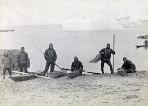 Seks menn står ved kajakker på en strand. Isfjell i bakgrunnen