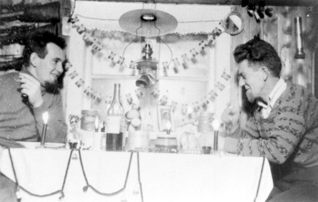 To menn sitter ved en bord med glass og flaskr. De har flippskjegg og ullgenser. Det henger julepynt foran vinduet
