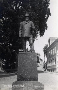 Statue av mann med hatt og skjegg i en gågate med en bygning på en side og trær på den andre