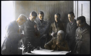 Seks menn står rundt en mann som sitter ved et skrivebord med et kart utlagt. De har uniform på seg og røyker pipe