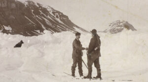 To menn handhilser i snøen med ski på bena. Til venstre en sort hund