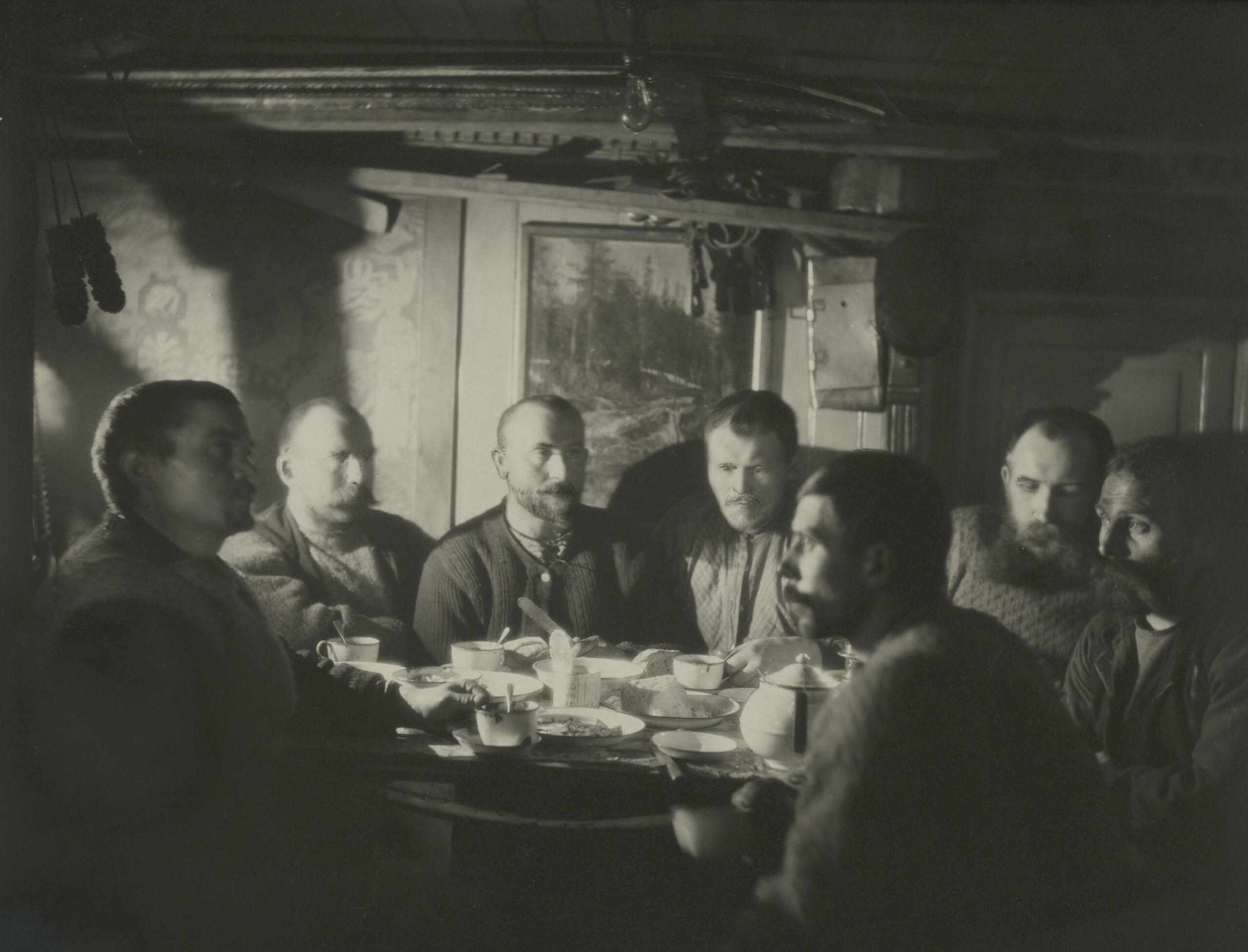 Syv menn sitter tett rundt et bord i et lite rom. De har ullklær og skjegg/bart.