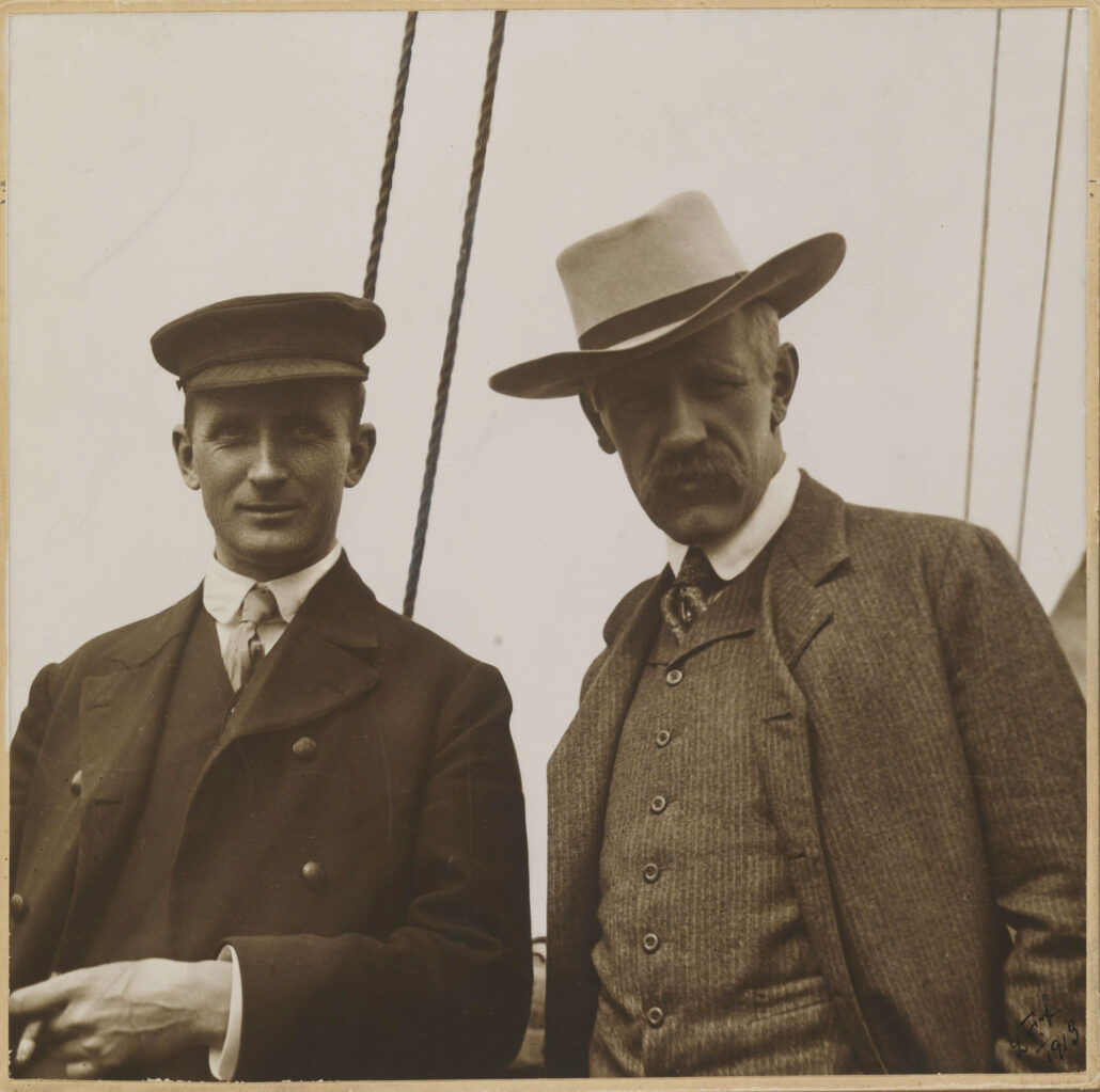 To menn med hatt og dress posererr på båtdekk
