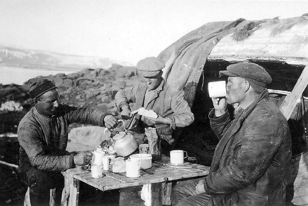 Tre menn spiser og drikker kaffe ved et enkelt trebord i fjæra, i bakgrunnen en veltet båt med presenning