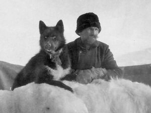 Mann med bart og vinterklær og en sort hund hviler armer/poter på et bjørnskinn som henger