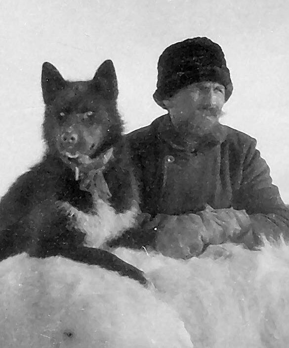 Mann med bart og vinterklær og en sort hund hviler armer/poter på et bjørnskinn som henger