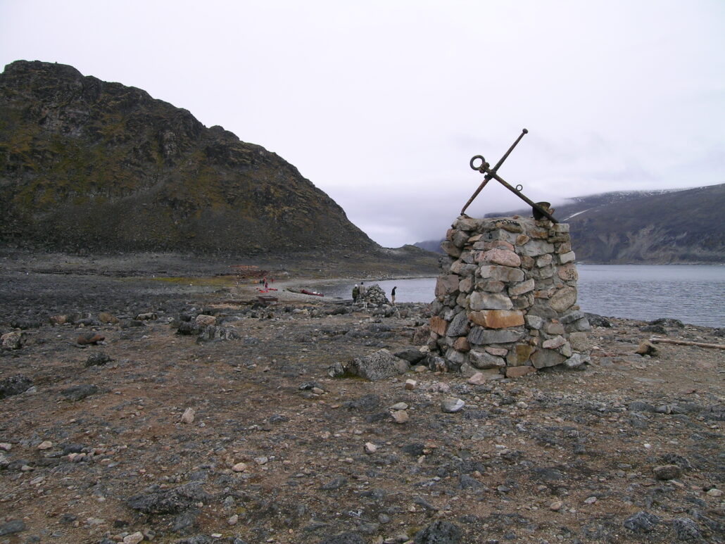 Stenvarde med et jernkors på toppen. Minnesmerket ligger på en strand og det er fjell i bakgrunnen.