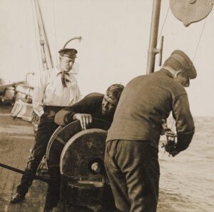 Mann i pene klær og hendene i bukselommen observerer to menn som arbeider på båtsdekk