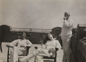 En mann og to kvinner slapper av på båtekk i pysjamaslignende hvite trøyer og bukser, kvinnene sitter i solstoler og mannen står og røyker.