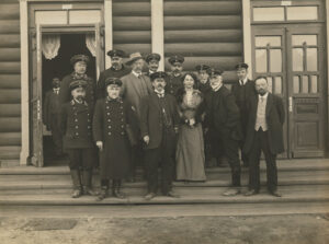 Gruppebilde av 14 personer på en trapp foran en laftet bygning. En kvinne i midten.