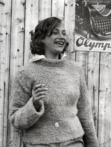Kvinne som røyker en sigarett. Hun har ullgenser på. På veggen bak ses en reklameplakat for Olympia skrivemaskiner