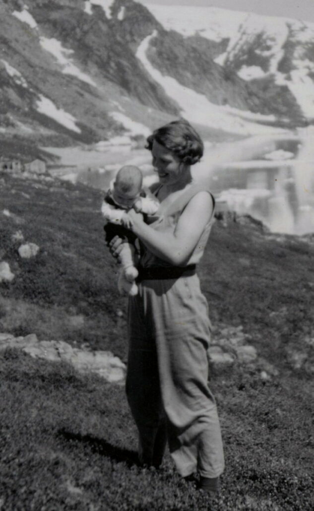 Kvinne holder et barn i armene, de står ved en fjord med snedekte fjell.