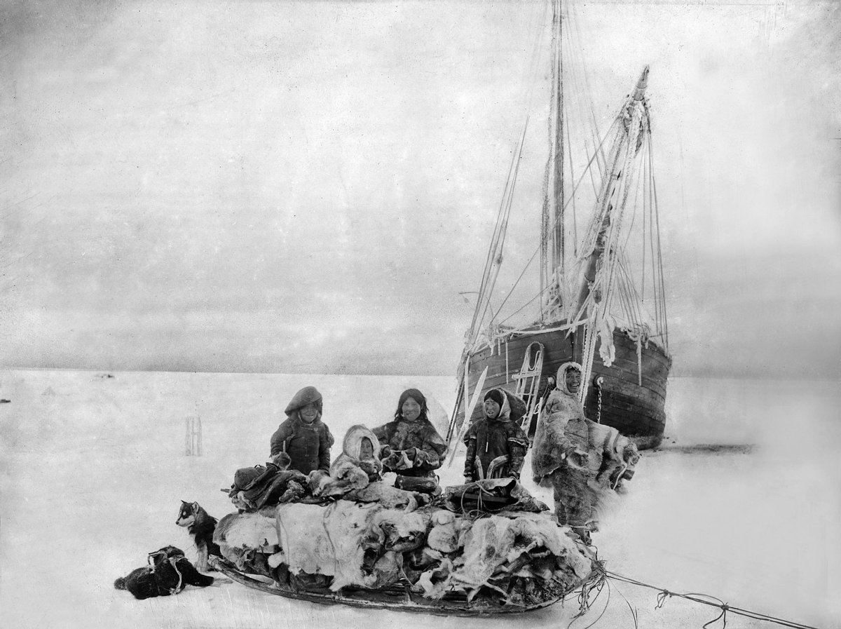 Fem inuitter i pelsklær står ved en fulllastet slede med en hund, i bakgrunnen en isdekket skute